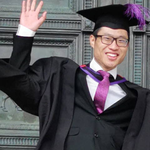 Man in graduation gown with mortar board, smiling with jazz hands